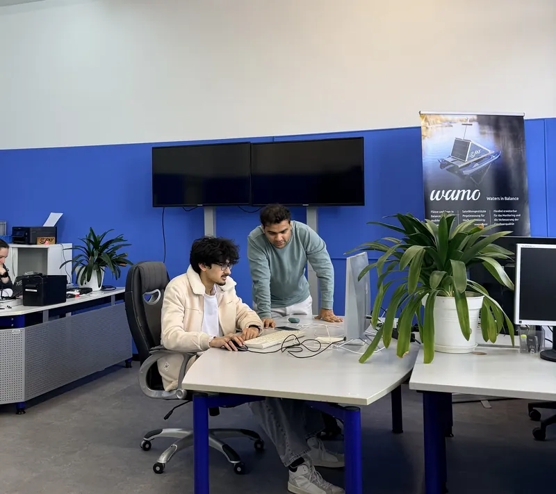 Zwei Kollegen vor dem Bildschirm im Büro in Darmstadt, einer sitzt am Rechner, der andere schaut mit