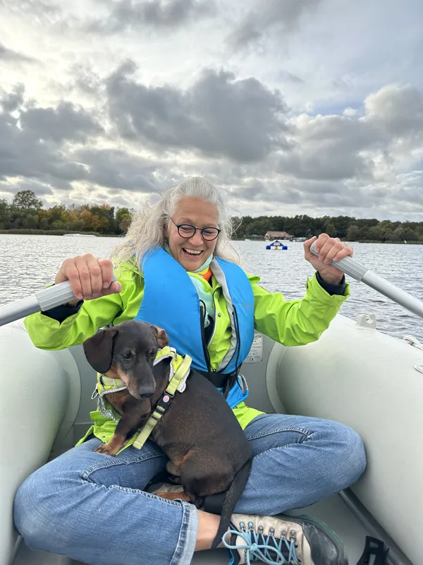 Iris Garbers lacht mit Dackel auf dem Schoß im Schlauchboot an den Rudern, WAMO-Station im Hintergrund, Mecklenburgische Seenplatte, Oktober 2024