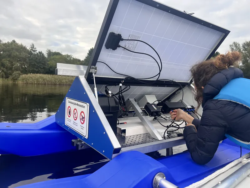 Kollegin arbeitet vom Boot aus an der Elektronik einer WAMO-Station, Solardeckel geöffnet, Mecklenburgische Seenplatte